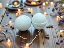 Load image into Gallery viewer, Two bath bombs on a wooden surface with decorative lights and herbs.