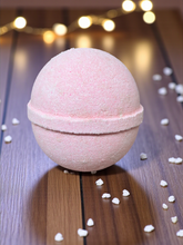 Load image into Gallery viewer, Pink bath bomb on a wooden surface with blurred lights in the background