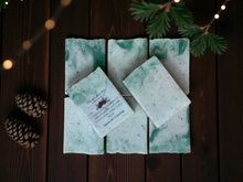 Load image into Gallery viewer, Handmade soap bars with a card on a wooden surface with decorative lights and pine branches.