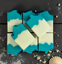 Load image into Gallery viewer, Handmade Seaside Cotton Soap bars with ocean-blue and cream layers, topped with sea salt-like crystals, displayed on a wooden surface with seashells and sand.