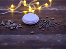 Load image into Gallery viewer, Purple bath bomb with small stones on a wooden surface with string lights in the background