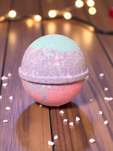 Load image into Gallery viewer, Multicolored bath bomb on a wooden surface with decorative lights in the background
