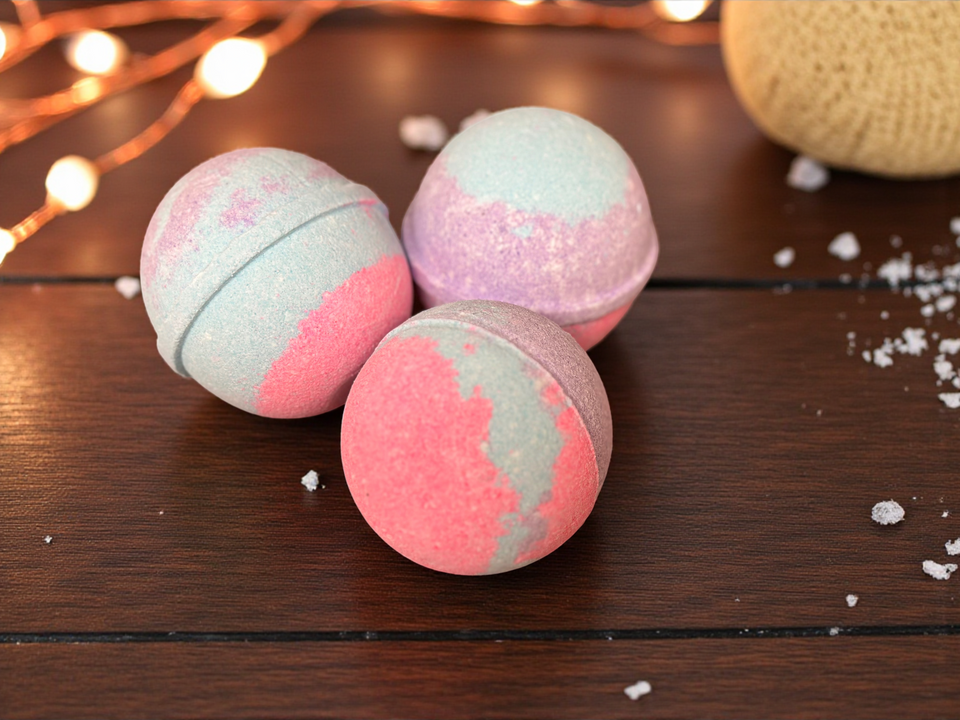 Three pink and gray bath bombs on a wooden surface with string lights in the background.