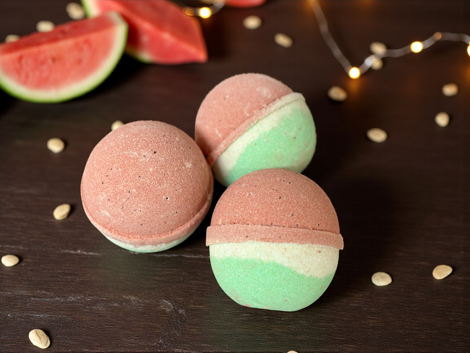 Two-layered pink and green bath bombs on a dark surface with watermelon slices in the background.